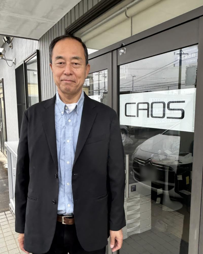 「CAOS」のサインがある入口の前に立つ、代表取締役の土岐昌彦
